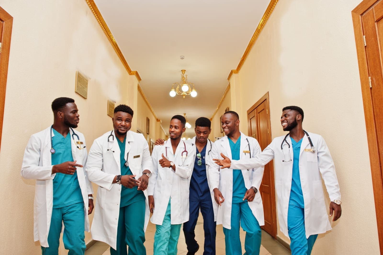 A group of clinical students in white coats and scrubs, stethoscopes around their necks, walking together and laughing down a hospital corridor